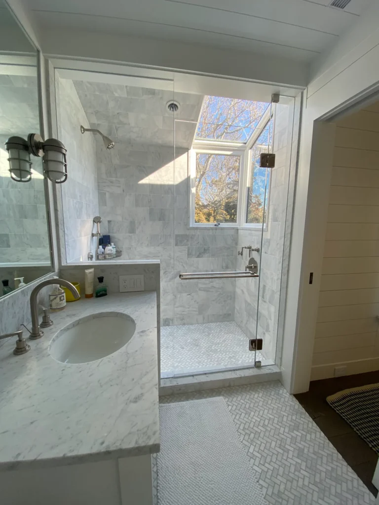A luxurious marble-tiled shower with a frameless glass door and a large overhead window providing natural light.