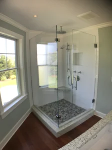 A frameless glass shower enclosure with a stone pebble floor, white tile walls, and a corner seat, set against a dark wood floor.