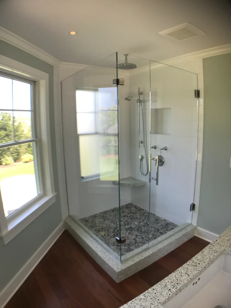 A frameless glass shower enclosure with a stone pebble floor, white tile walls, and a corner seat, set against a dark wood floor.
