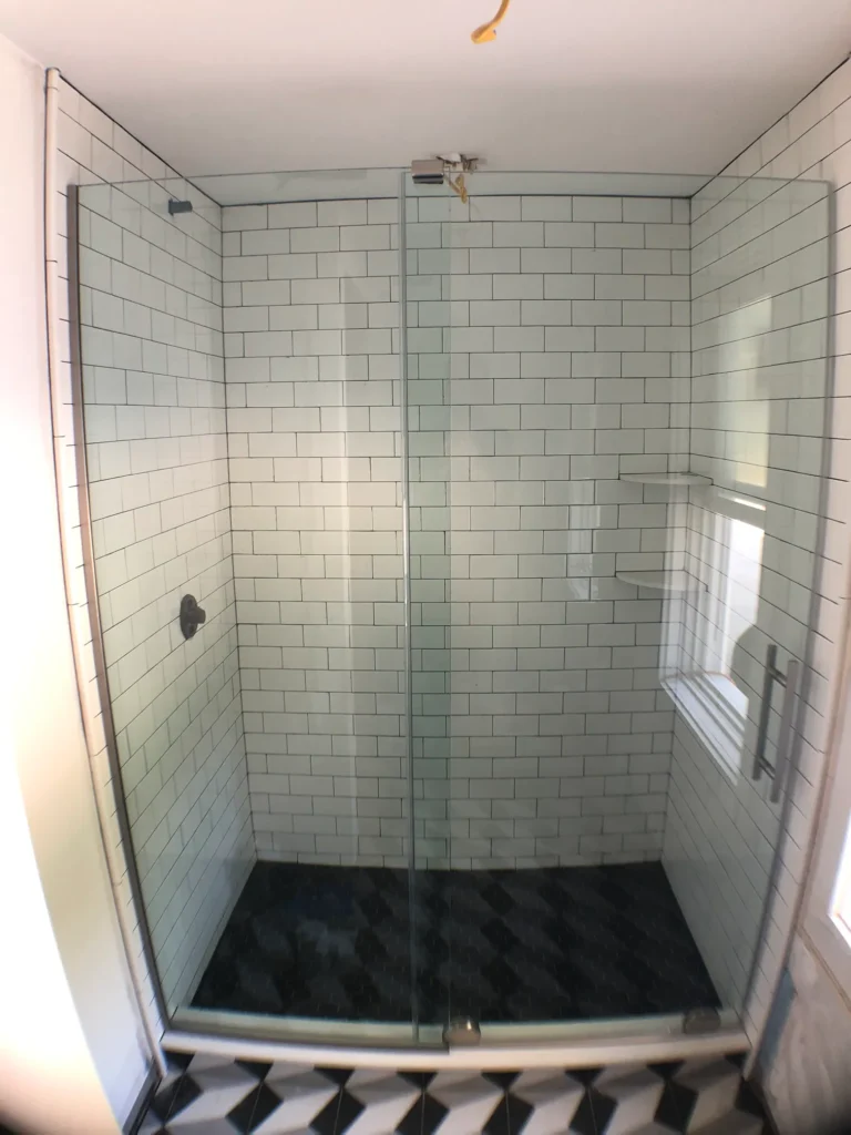 A frameless glass shower enclosure featuring white subway tile walls and a contrasting black patterned floor.