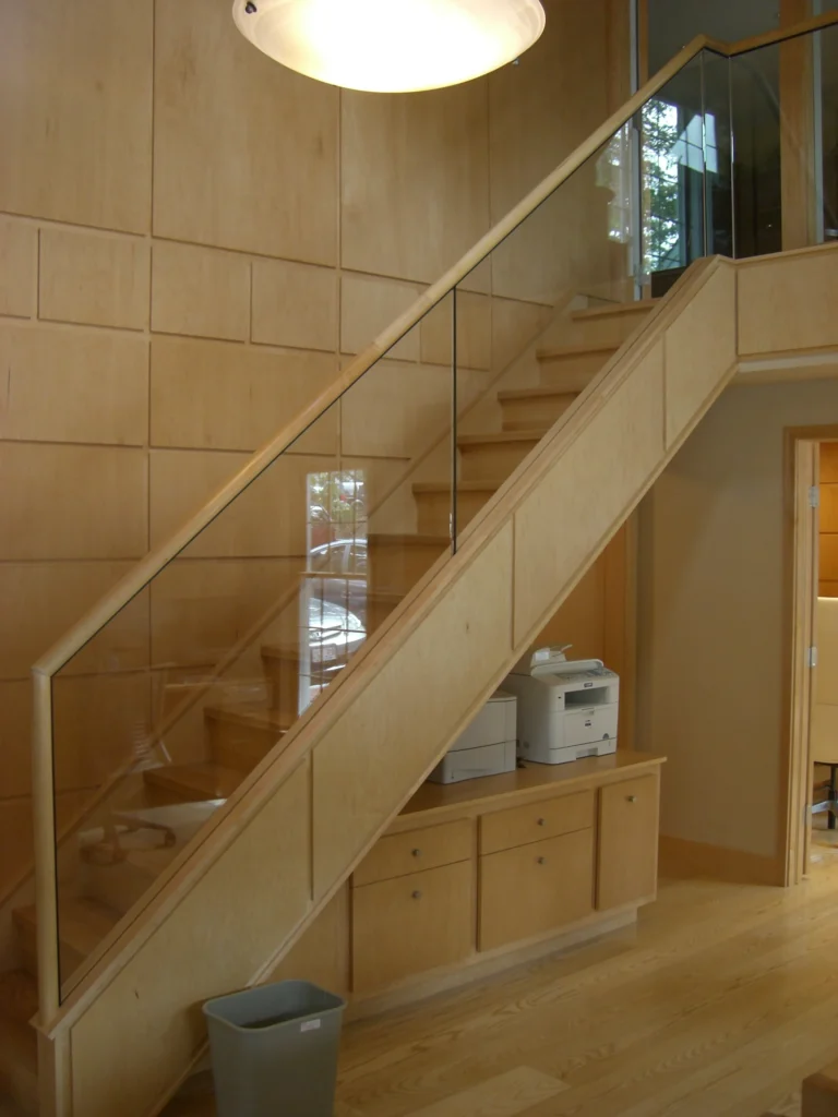 A light wood staircase and matching wall paneling featuring a sleek glass railing and a small built-in storage unit beneath.