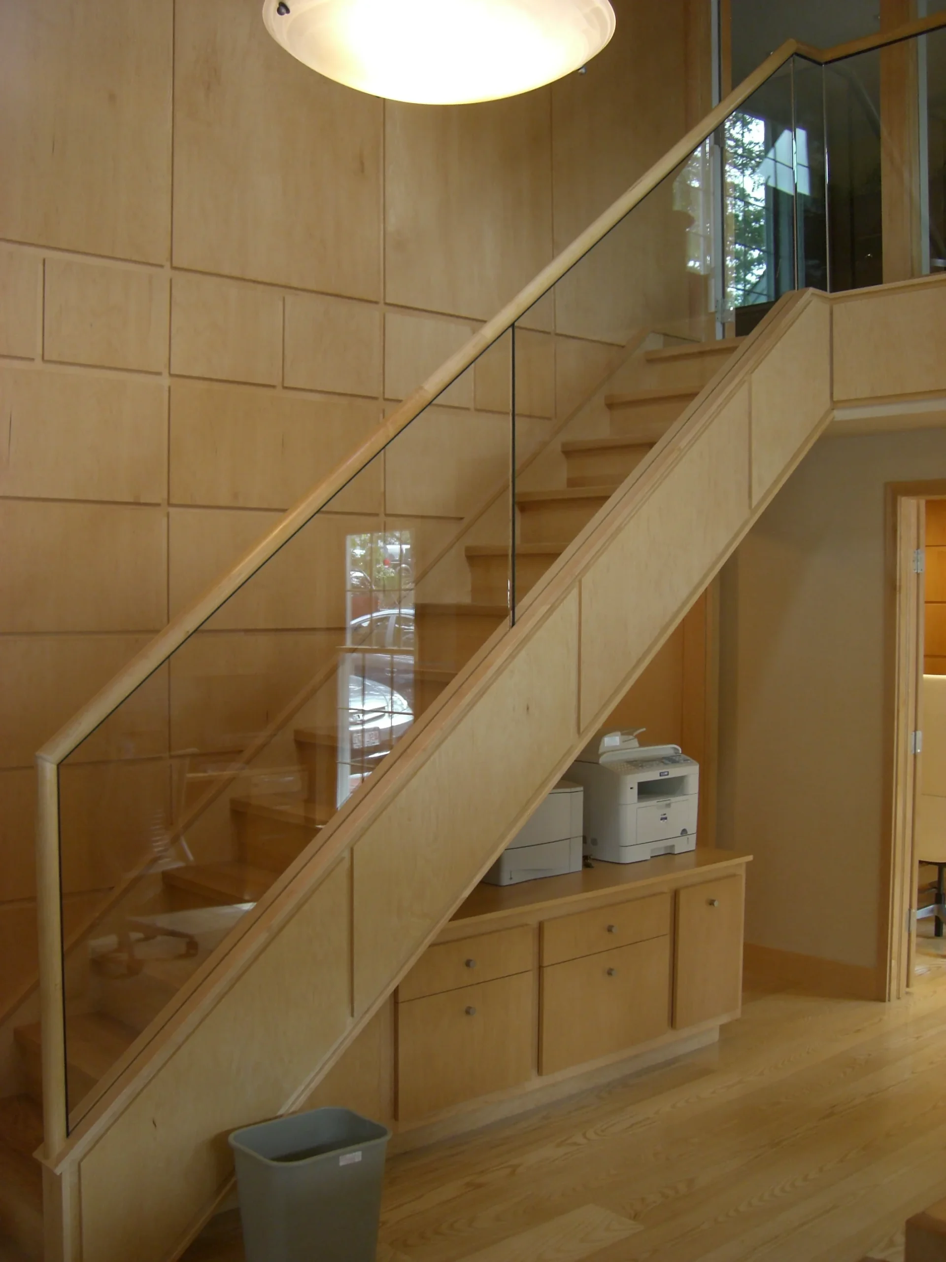 A light wood staircase and matching wall paneling featuring a sleek glass railing and a small built-in storage unit beneath.