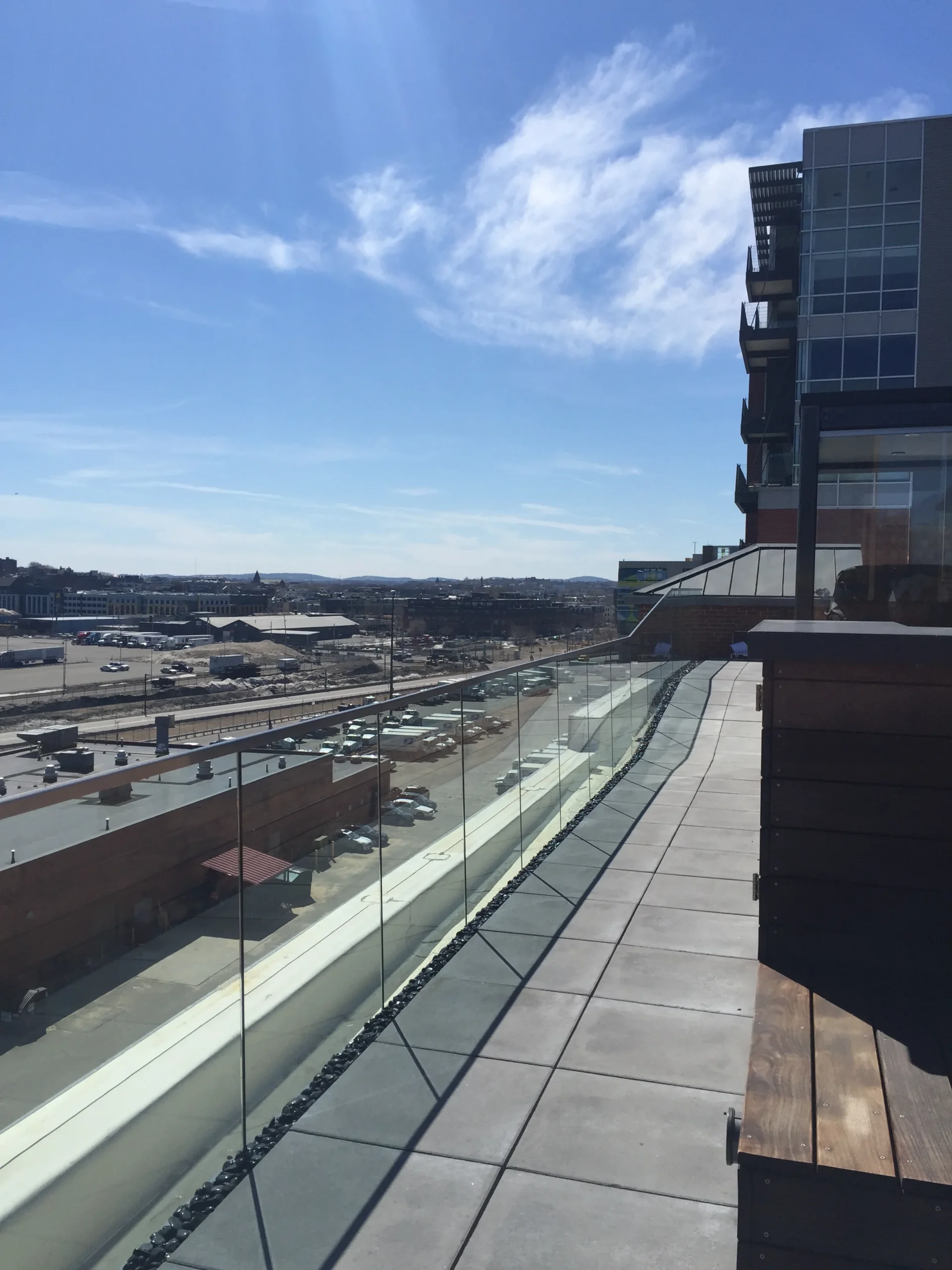 A grey-tiled rooftop balcony with a frameless glass railing overlooking a city industrial area under a bright blue sky.