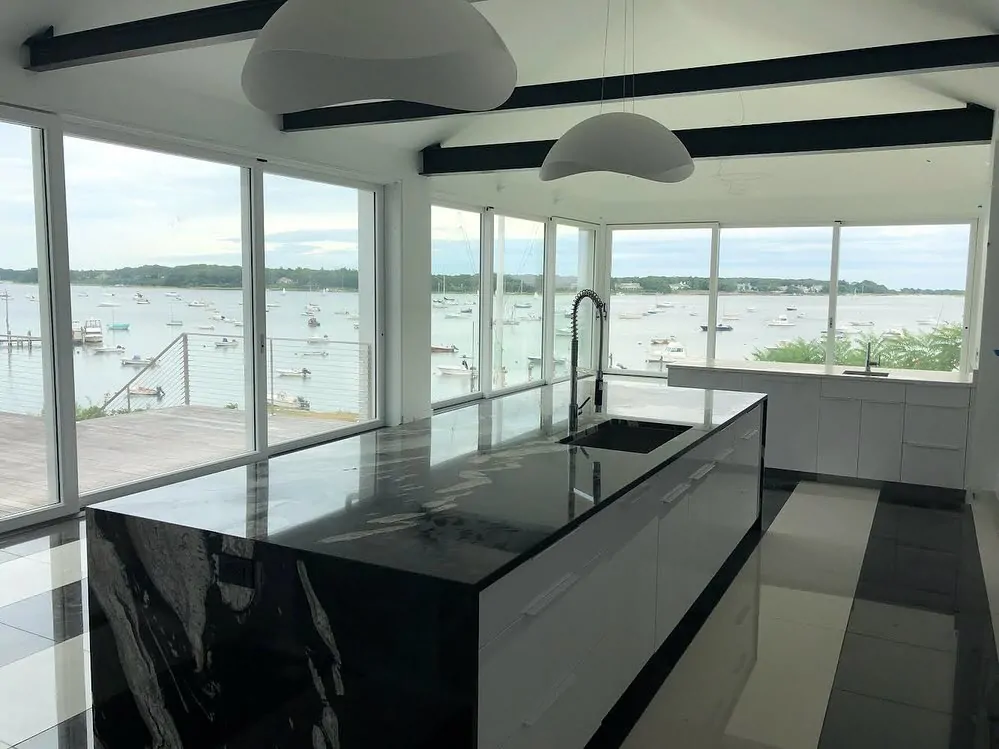 A dark marble kitchen island in a bright white room with large windows overlooking a harbor full of boats.