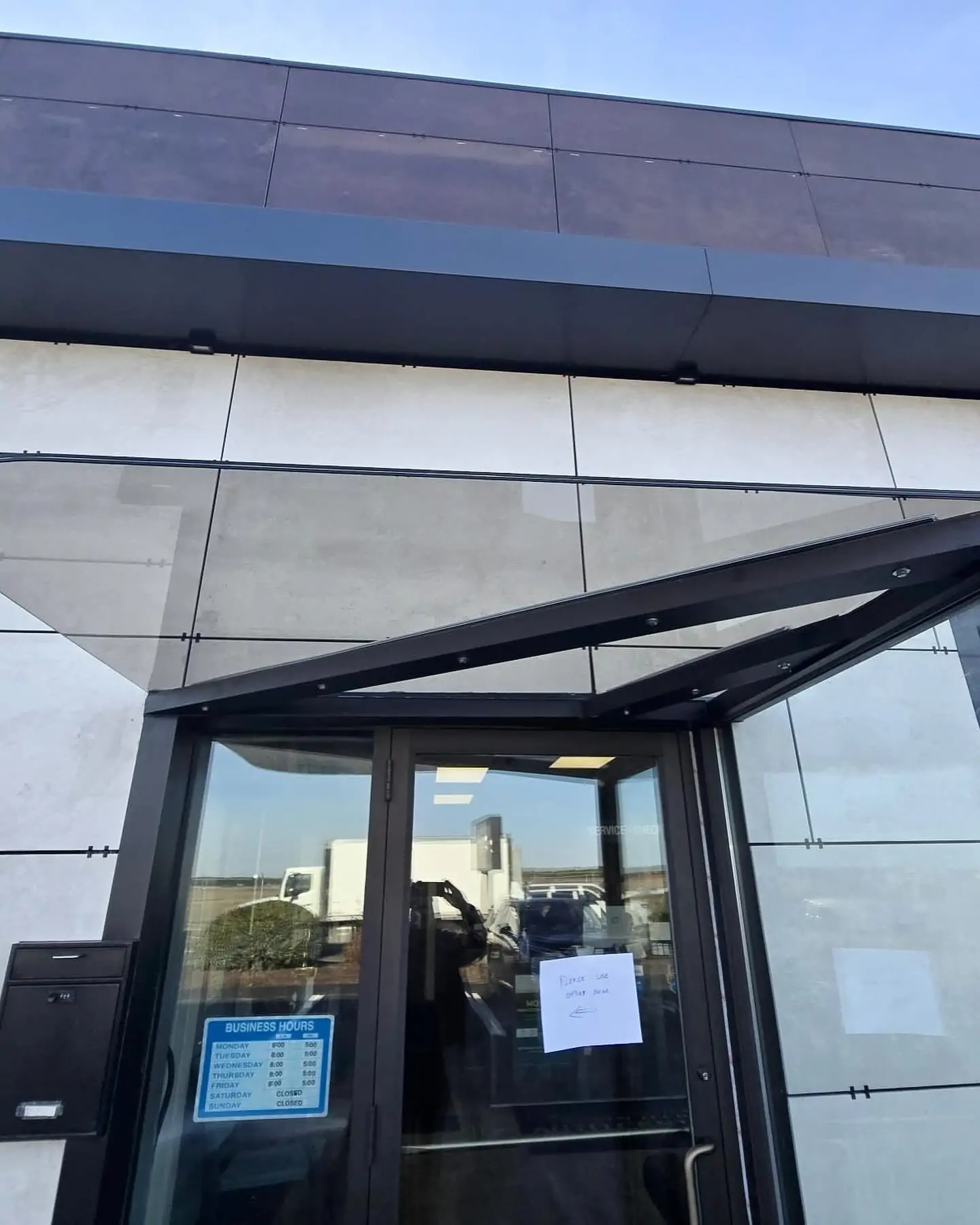 A dark-framed glass door under a glass awning, featuring a "Business Hours" sign and a white note taped to the glass.